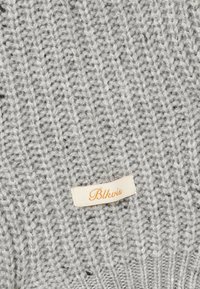 Tissu tricoté gris doux avec une petite étiquette en tissu crème cousue, comprenant un texte en cursive orange indiquant "Blkvis."