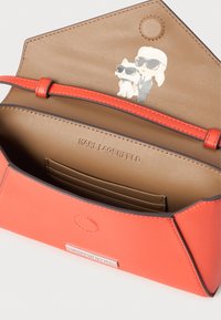 Geantă din piele roșie cu interior bej, având sloturi pentru carduri și un grafic de desene animate cu o figură și o pisică purtând ochelari de soare. Logo-ul este embossat în interior.