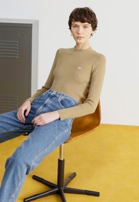 Gerippter, hellbrauner Langarm-Oberteil mit einem Stehkragen, kombiniert mit hochgeschnittenen blauen Jeans. Das Model sitzt auf einem Holzstuhl vor einem gelben Boden.