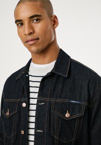 Chaqueta de mezclilla oscura con botones de cobre, costuras en contraste, dos bolsillos en el pecho, llevada sobre una camisa a rayas blanca y navy.