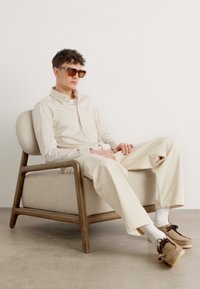 Camicetta a righe beige, pantaloni chiari e scarpe marroni su una persona seduta in una sedia a sdraio curva di legno con un cuscino di stoffa.
