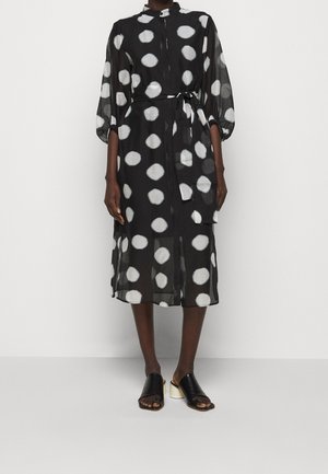 Vestido midi negro con grandes lunares blancos, mangas abullonadas, cinturón en la cintura, llevado con sandalias negras de tacón bloque y punta abierta.