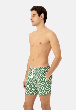 Nuori mies, jolla on lyhyet tummat hiukset, pukeutuneena vihreisiin kuvioituihin uimashortseihin, joissa on valkoinen nyörikiristys, seisoo kädet taskuissa vasemmalle päin.