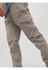 Person wearing beige cargo pants with flap pockets on thighs, standing against a plain light background.