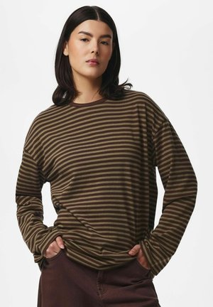 Jeune femme aux cheveux foncés mi-longs portant un chemisier à manches longues rayé marron et vert, les mains dans les poches.