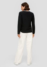 Schwarzer langärmeliger Strickpullover mit strukturiertem Finish, kombiniert mit lockeren weißen Hosen und flachen weißen Sneakern.