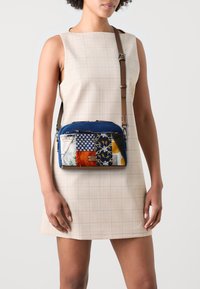 Modrá kabelka přes rameno s různými vzory a texturami tkanin, vybavená zipem a nastavitelným hnědým popruhem.