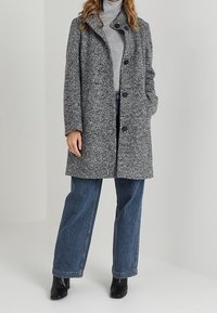 Manteau gris texturé avec un col haut, des boutons de fermeture et des poches latérales, porté sur un léger pull et un jean bleu à jambe large. Bottes noires à la cheville.