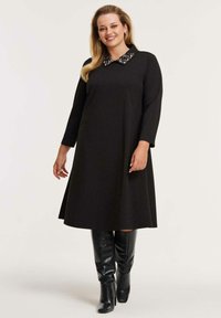 Schwarzes Kleid mit langen Ärmeln und einem mit Schmucksteinen verzierten Kragen, mit einem ausgestellten Rock und einer glatten Textur, kombiniert mit glänzenden kniehohen Stiefeln.