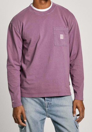 Muž nosící vybledlou fialovou košili s dlouhým rukávem a kapsou na hrudi, světlé modré cargo džíny, stojící s rukama volně podél těla.