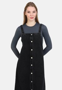 Abito in denim nero con spalline regolabili, chiusura frontale a bottoni e tasche laterali. Il modello indossa una maglietta grigia a maniche lunghe sotto.