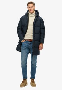 Abrigo puffer azul marino con capucha, diseño acolchado, combinado con un suéter de cuello alto beige, jeans azules y botas marrón claro. Presenta un cierre de cremallera y botón.