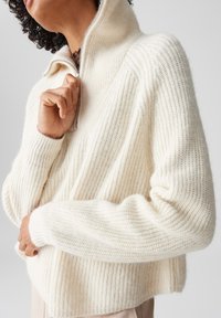 Maglione lavorato a maglia color crema con alto colletto e mezza zip. Presenta una texture a coste e spalle scese. Design morbido e spesso.