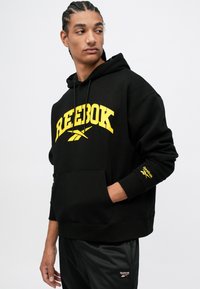 Čierna mikina s kapucňou a veľkým žltým logom Reebok, kengurská kapsa a akcentované logo na rukáve. Mäkký materiál a voľný strih. Zodpovedajúce čierne nohavice.