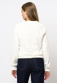 Vrouw met lang lichtbruin haar, gekleed in een witte lange mouwen shirt en donkerblauwe jeans, van achteren gezien tegen een witte achtergrond.