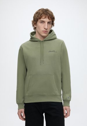 Jeune homme aux cheveux châtain clair portant un sweat à capuche Lacoste vert olive avec une poche avant et un pantalon noir, debout devant un fond uni.