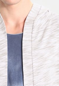 Lätt, texturerad stickad cardigan i beige med subtilt zigzagmönster, lagd över en enfärgad blå topp, med en öppen framsida.