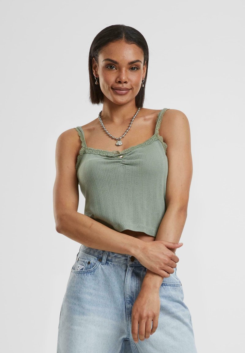 Groen geribbeld crop-top met kantafwerking, met een knoopdetail in het midden. Draag met lichtblauwe denim jeans.