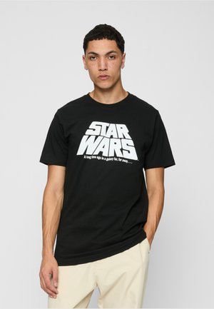 Junger Mann mit kurzen, lockigen Haaren, der ein schwarzes "Star Wars"-T-Shirt und beige Hosen trägt, vor einem schlichten hellen Hintergrund steht.