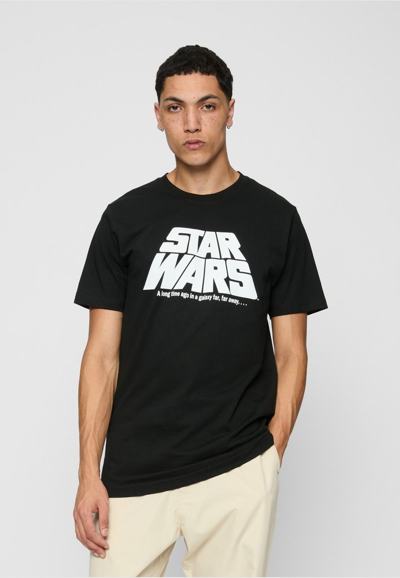 Jeune homme aux cheveux courts et bouclés portant un t-shirt noir « Star Wars » et un pantalon beige, debout devant un fond clair uni.