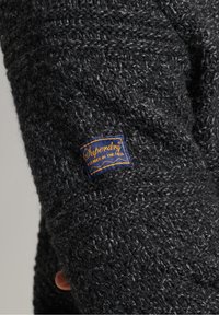 Gestreifter Pullover in Dunkelgrau mit einem strukturierten Muster. Verfügt über ein blaues und goldenes "Superdry"-Etikett am rechten Ärmel.