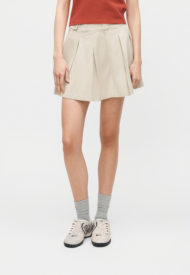 Donna con minigonna plissettata beige, calzini grigi e sneakers bianche con lacci e disegni neri, in piedi contro uno sfondo semplice.