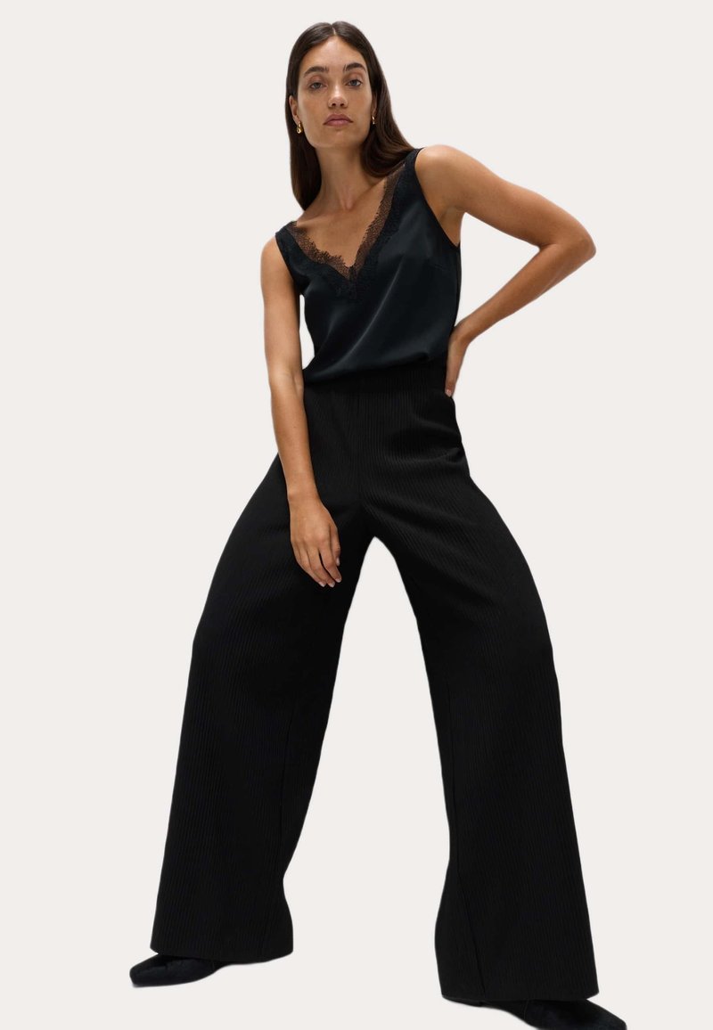 Camisón de satén negro con borde de encaje, combinado con pantalones de pierna ancha plisados en negro. Las telas presentan un contraste suave y texturizado.