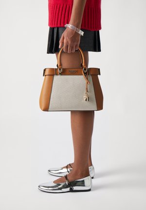 Sac à main structuré beige et fauve en toile et cuir, avec des éléments en métal doré et un pendentif. Le modèle porte des ballerines argentées et un pull rouge.