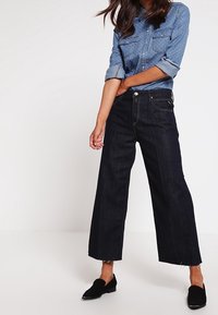 Donna che indossa una camicia a pulsante blu a fantasia con le maniche arrotolate, jeans larghi scuri e scarpe slip-on nere su uno sfondo semplice.