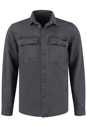 Mężczyzna idący w szarej zapinanej na guziki kurtce, zielonym swetrze, zniszczonych niebieskich jeansach i czarno-białych tenisówkach na białym tle.