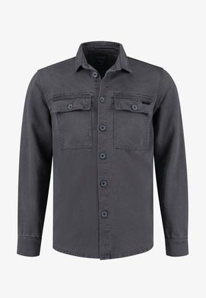 Mann, der geht, trägt eine graue, geknöpfte Jacke, grünen Pullover, zerrissene blaue Jeans und schwarz-weiße Turnschuhe vor weißem Hintergrund.