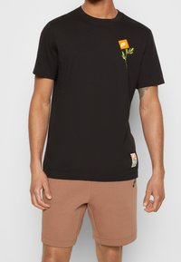 Svart bomullströja med en grön bladdesign och en orange Nike-logotyp på bröstet. Visas med bruna shorts.