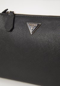 Černá texturovaná zipová taška s trojúhelníkovým kovovým logem Guess USA a koženým táhlem na zip na bílém pozadí.