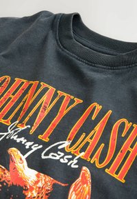 Černá mikina s žebrovaným límcem. Obsahuje výrazný žlutý a červený text "JOHNNY CASH" a bílý podpis s grafikou orla. Viditelná opotřebovaná textura.