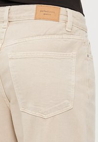 Ljusbeiga jeans med en jämn yta, som har en tan läderetikett med texten "perfekta jeans" och en enda bakficka med sömnadsdetaljer.