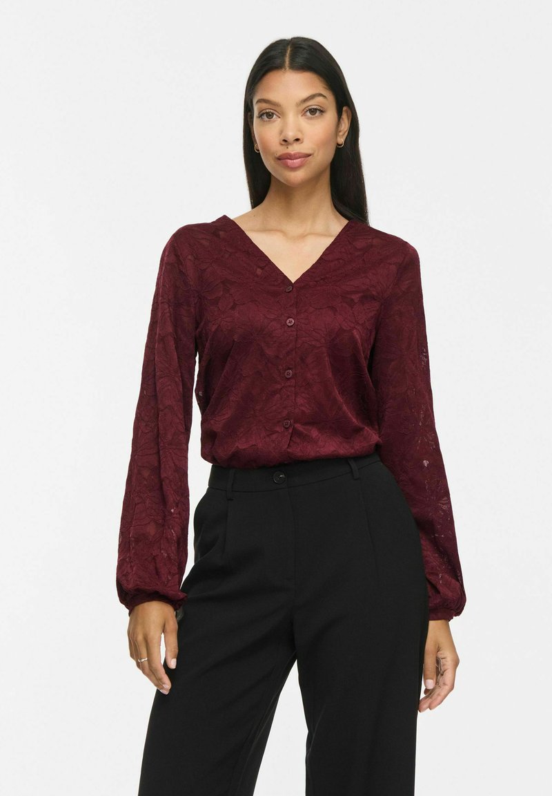 Femme aux longs cheveux foncés portant un chemisier en dentelle bordeaux boutonné et un pantalon noir taille haute, debout sur un fond uni clair.