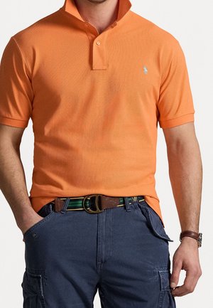 Oranje poloshirt met een gekraagde hals en een tweeknopssluiting, gecombineerd met donkere cargoshorts. Bruine riem met gouden gesp en groene strepen.