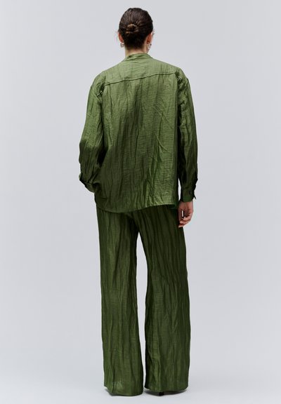 Personne aux cheveux bruns en chignon portant une chemise ample et texturée à manches longues vert olive et un pantalon large assorti, debout dos à la caméra.