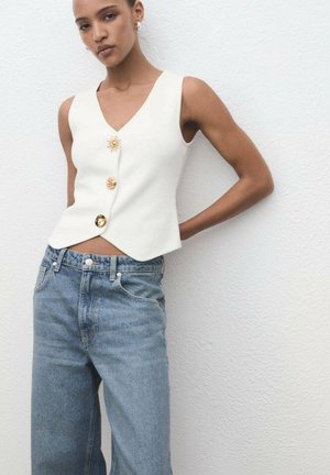 White sleeveless top with a V-neck and angled hem, featuring three gold buttons and a sun-shaped accent, paired with light blue jeans.