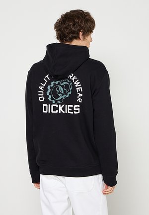 Personne portant un sweat à capuche noir avec un logo d'engrenage bleu et un texte blanc "QUALITY WORKWEAR DICKIES" dans le dos, associé à un pantalon blanc.