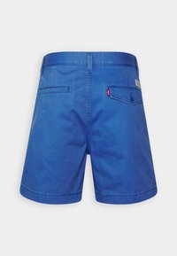 Pantalones cortos de mezclilla azul con una textura suave, que presentan dos bolsillos traseros, un cierre de botón y una pequeña etiqueta de logo roja en la cinturilla.