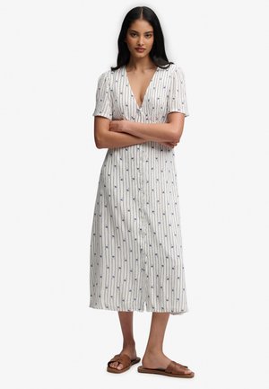 Vrouw met lang zwart haar die een witte midi-jurk draagt met blauwe verticale strepen en bloemenprint, bruine slidesandalen, staand met gekruiste armen.