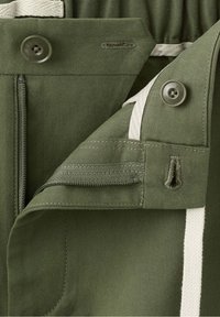 Ceinture en tissu vert avec braguette zippée, fermeture par bouton, doublure intérieure beige et boutonnières visibles sur un pantalon.