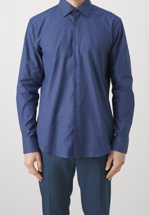 Homme portant une chemise bleu marine cintrée à manches longues, boutonnée avec col, et un pantalon bleu marine assorti, debout sur un fond uni.