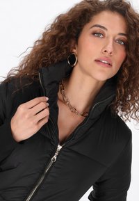 Fekete puff kabát magas gallérral, elülső cipzárral és bordázott mandzsettákkal. Arany színű kiegészítők és egy láncnyaklánc részletezi.