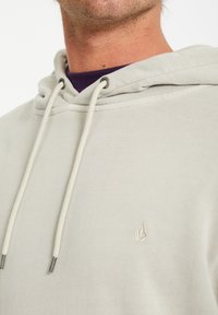 Lys beige hoodie med snore, der har et lille broderet logo på brystet og en tekstureret stoffinish.