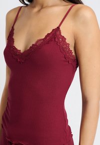 Burgundy camiset med ribbat mönster, spetsdetaljer längs V-halsen och tunna justerbara axelremmar. Har en figurnära silhuett.