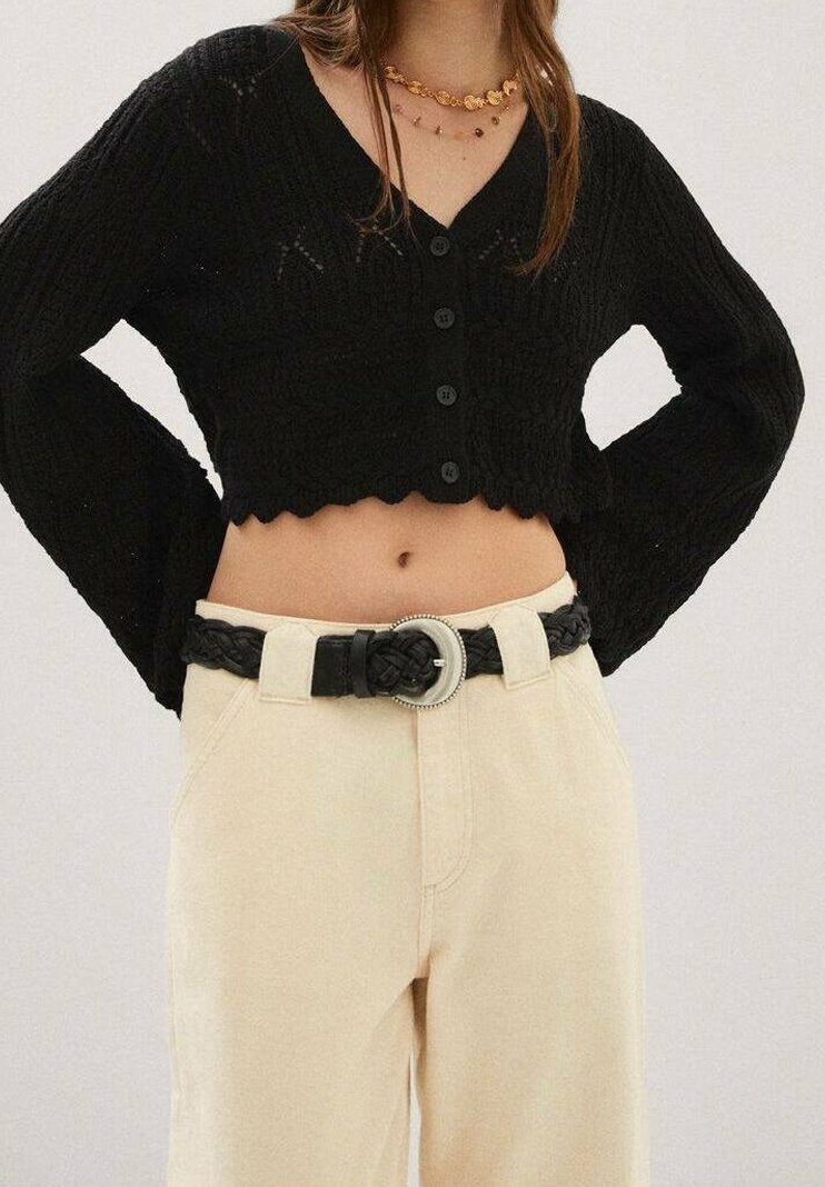 Zwarte gebreide cropped cardigan met een golvende zoom en knoopsluiting aan de voorzijde, gecombineerd met beige high-waisted broeken en een zwarte gevlochten riem.