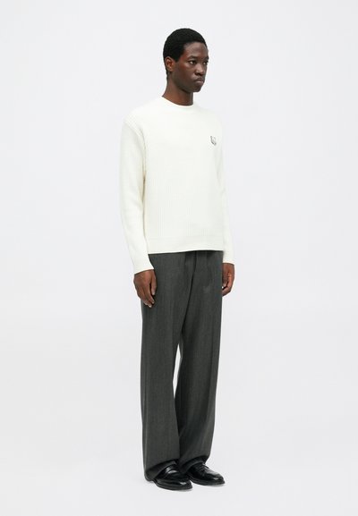 Man wearing a white ribbed sweater with a small emblem, loose gray trousers, and black shoes, standing against a plain white background.