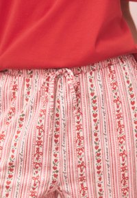 Zbliżenie na czerwony top włożony w białe spodnie z czerwonym i zielonym kwiatowym oraz sercowym wzorem i sznurkiem związanym z przodu.